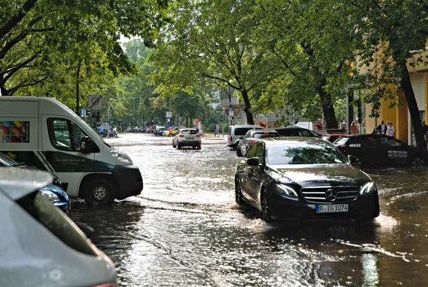 Überschwemmte Straße in Berlin, Deutschland mit Autos, die durch das Wasser fahren, Fußgängern auf dem Gehweg, Bäumen entlang der Straße, Gebäuden im Hintergrund und teilweise untergetauchten Schildern und Polen.