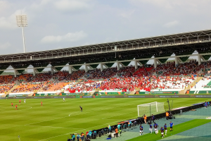 Ein Fußballspiel in einem großen Stadion mit Spielern auf dem Boden, einem Tor, einem Zaun, einer Fahne mit Text, Metallrahmen, einem Dach mit Deckenleuchten und einem bewölkten Himmel.