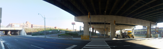 A truck driving under a bridge on a city street with buildings, street infrastructure, and vehicles visible under a clear blue sky.