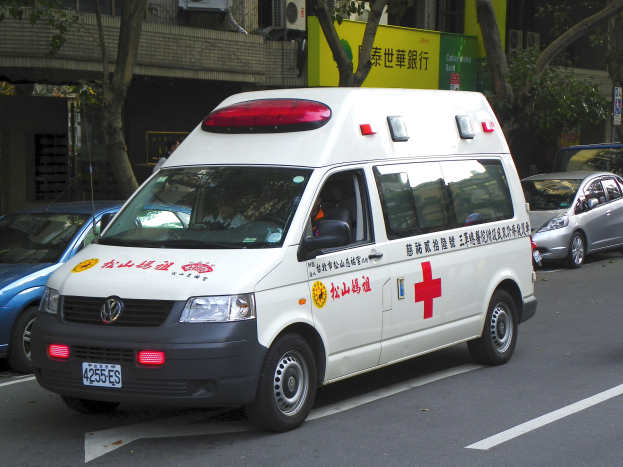 Ambulanz am Straßenrand geparkt mit anderen Fahrzeugen, Bäumen, Gebäuden, Schildern und Klimaanlagen im Hintergrund.