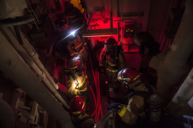 Feuerwehrleute in Helmen und Sauerstoffflaschen stehen in einem schwach beleuchteten Raum mit Rohren auf beiden Seiten und bereiten sich auf einen Rettungseinsatz in der Nähe von Maschinen und anderen Gegenständen vor.