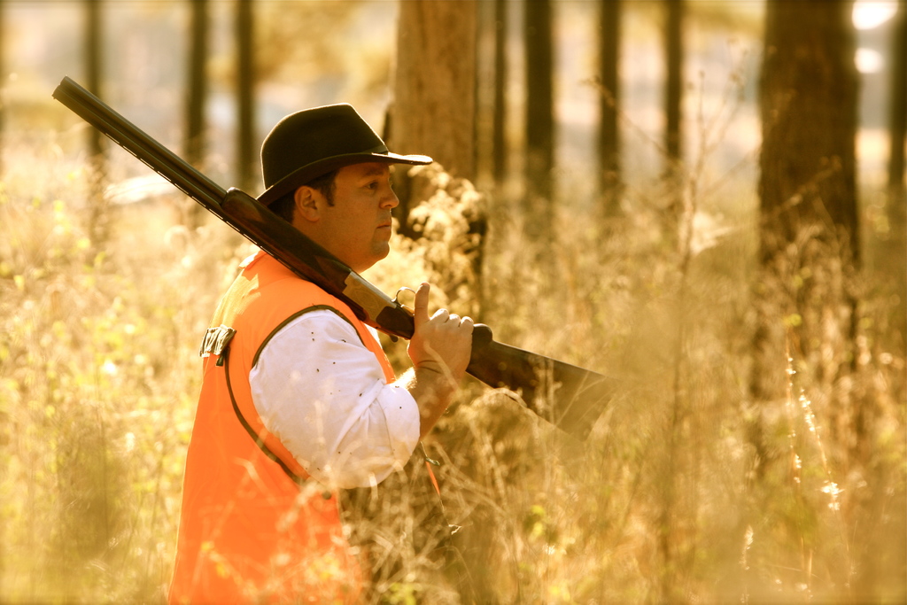 Eine Person mit einer Waffe steht vor Pflanzen, mit Bäumen im Hintergrund.