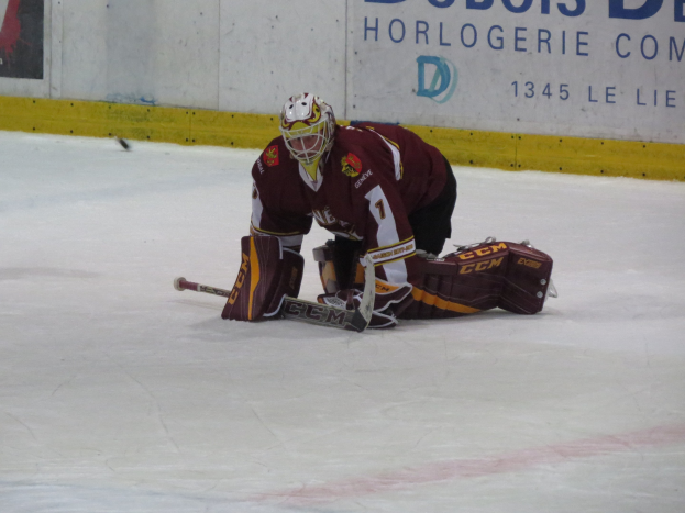 Eishockeyspieler in rot-gelber Uniform, der einen Schuss auf dem Eis abwehrt, trägt Helm, Handschuhe und Knieschoner, hält einen Eishockeyschläger und hat eine Wand und Text im Hintergrund.