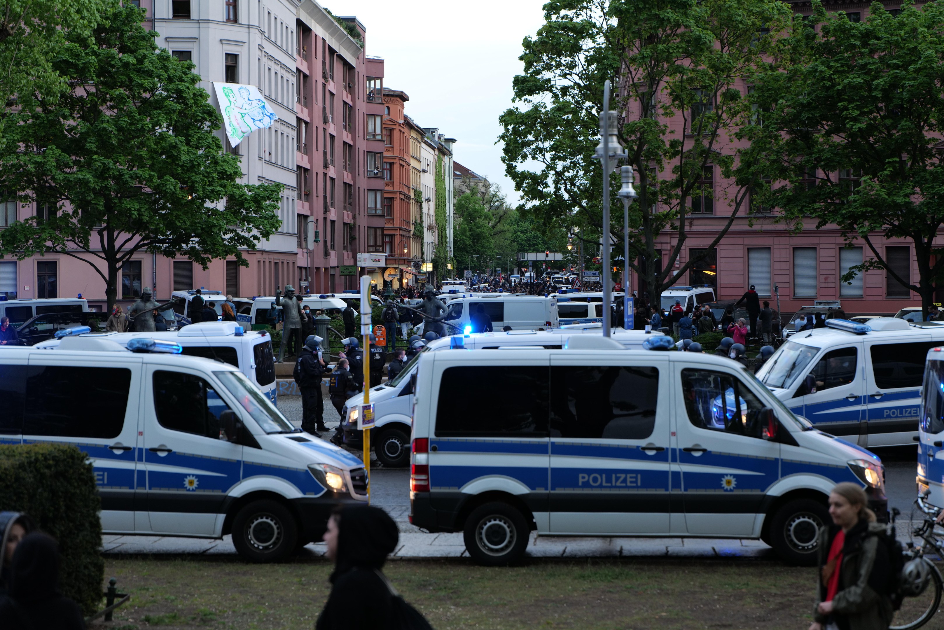 Gruppe von Polizeiwagen auf einer Straße geparkt mit Menschen und Fahrrädern in der Nähe, Bäume und Gebäude säumen die Straße, ein Banner im Hintergrund und sichtbarer Himmel.