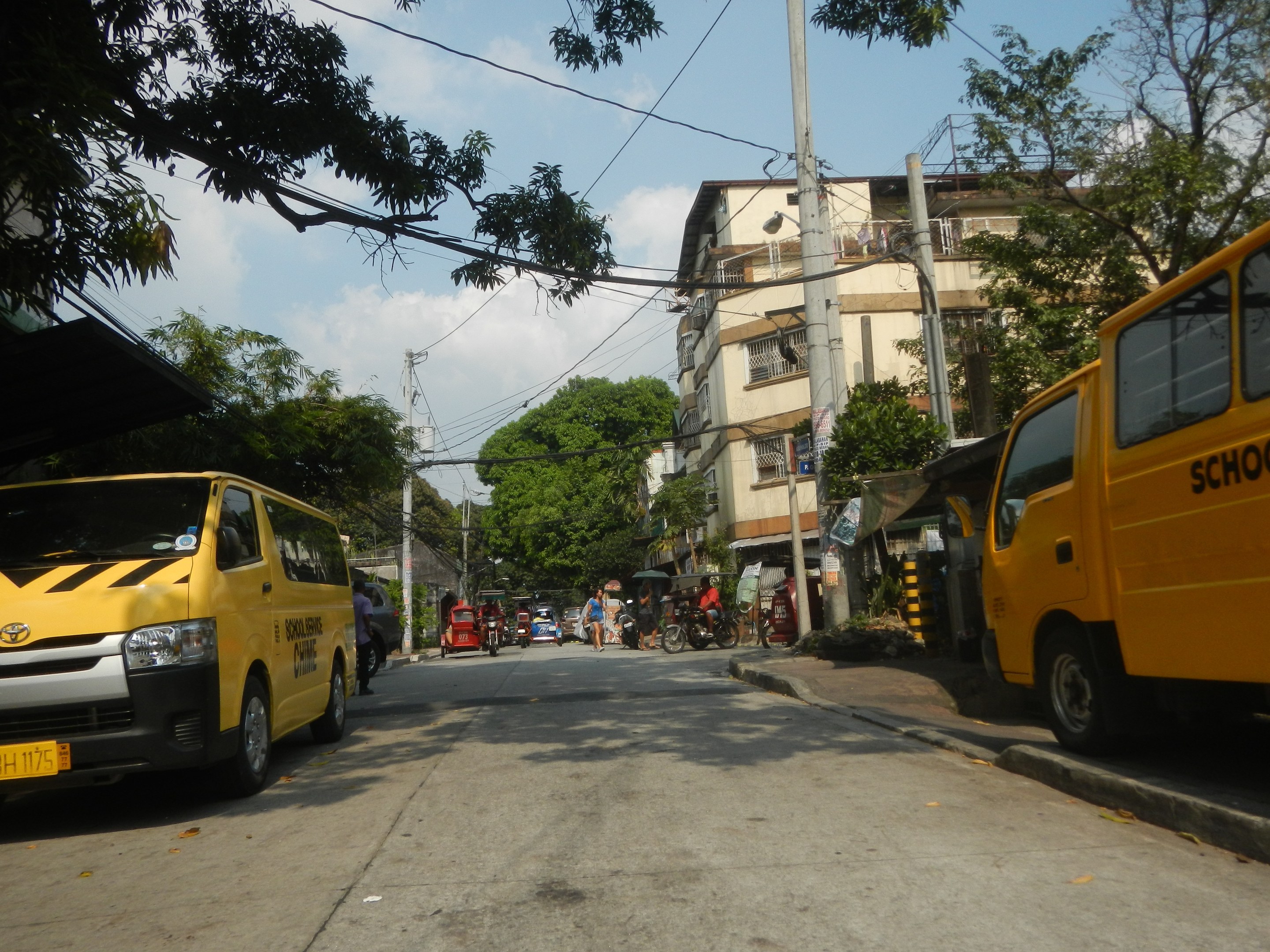 A yellow school bus travels down a street with tall buildings, trees, electric poles with wires, cyclists, and other vehicles under a cloudy sky.
