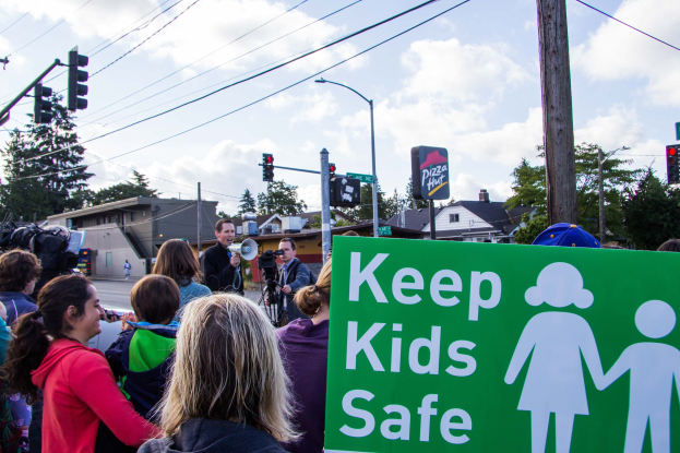 Gruppe von Menschen an einer Straßenseite mit einer "Keep Kids Safe"-Plakette, mit einem Mann im Vordergrund, der ein Megafon hält, und Gebäude, Bäume und Verkehrsinfrastruktur im Hintergrund.