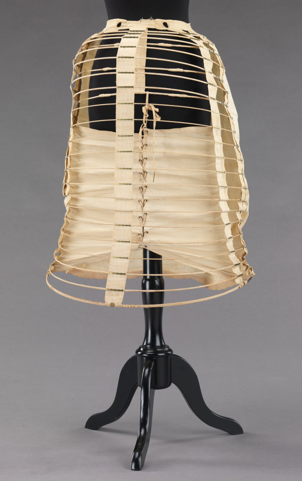 Eine Schaufensterpuppe trägt einen hellfarbenen, hochtaillierten, plissierten Rock mit einer gerüschten Saumkante über dem Knie auf einem schwarzen Ständer vor einem grauen Hintergrund.