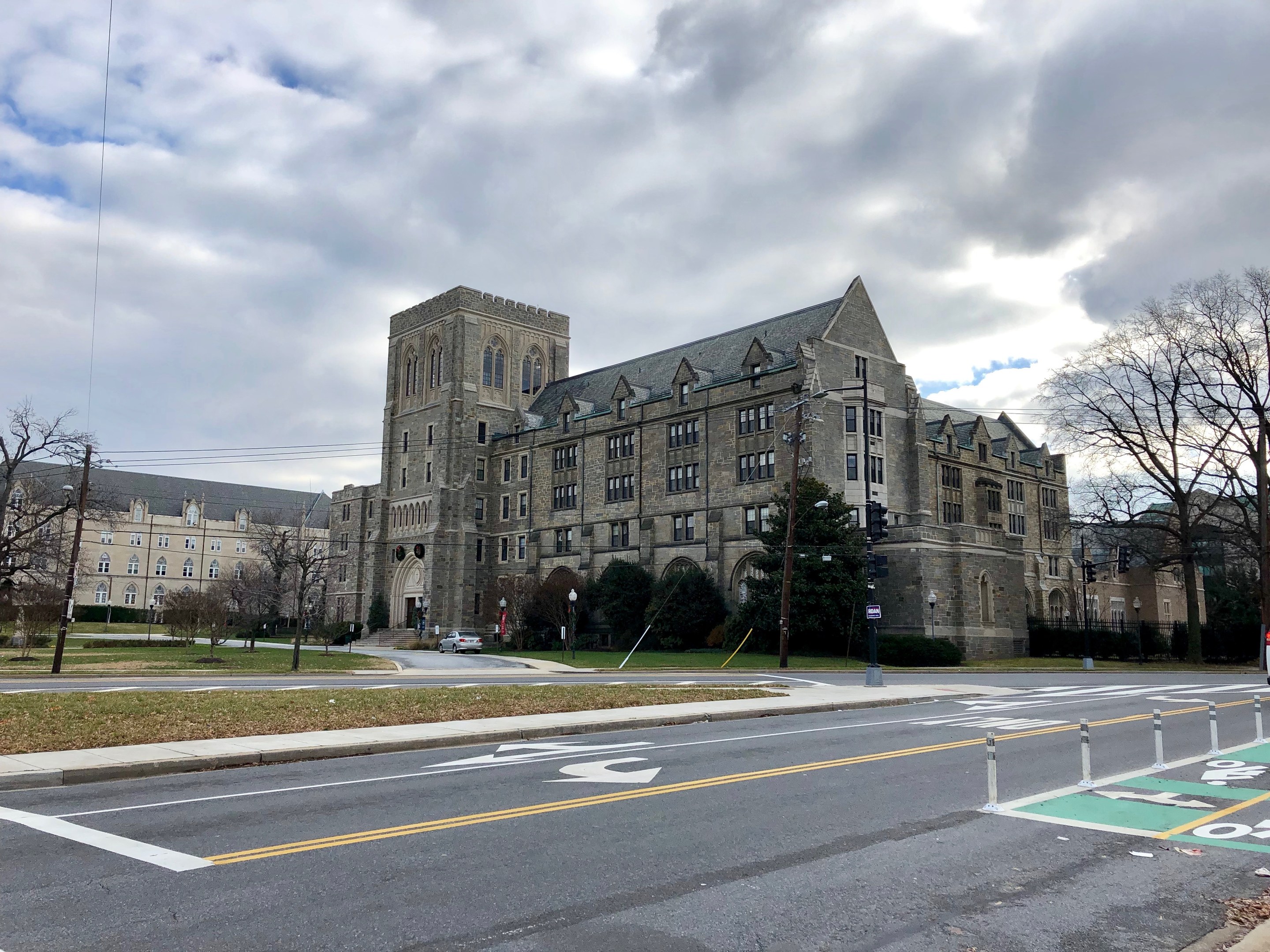 Großes Gebäude mit zahlreichen Fenstern, als der neue Campus der Universität Pennsylvania bezeichnet, umgeben von Bäumen, Straßenlaternen, Verkehrssignalen und Fahrzeugen unter einem bewölkten Himmel.