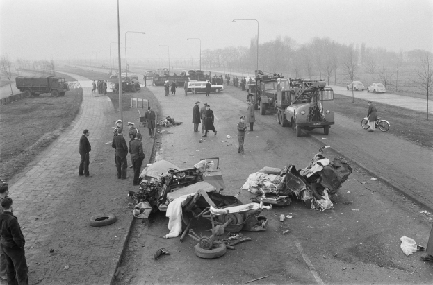 Schwarz-weißes Bild einer Autounfall-Szene am Straßenrand mit mehreren Fahrzeugen, Einsatzkräften und Schaulustigen, umgeben von Laternen, Bäumen und Himmel.