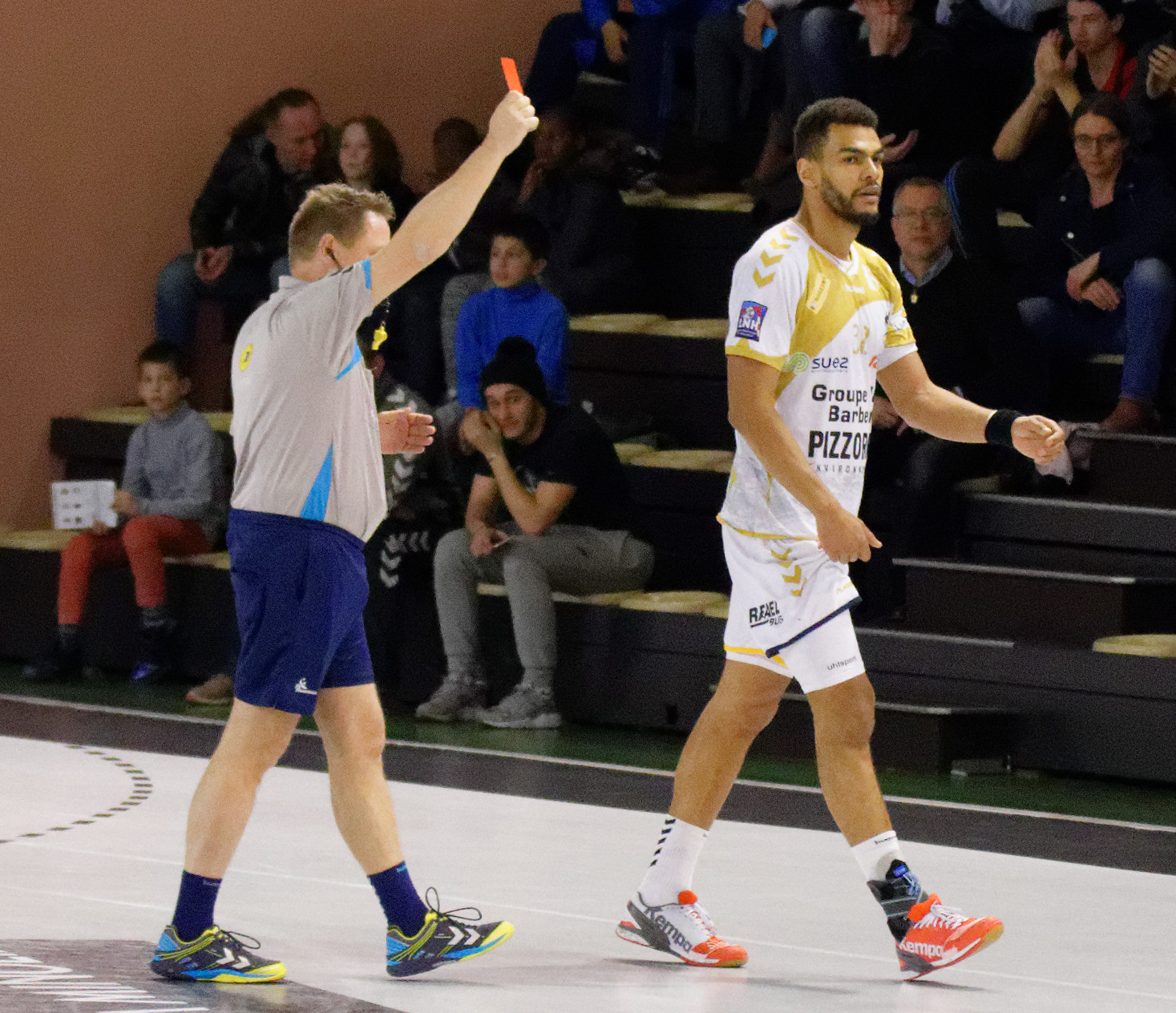 Zwei Männer in Sportuniformen spielen Handball vor einer sitzenden Menge mit einer Wand im Hintergrund.