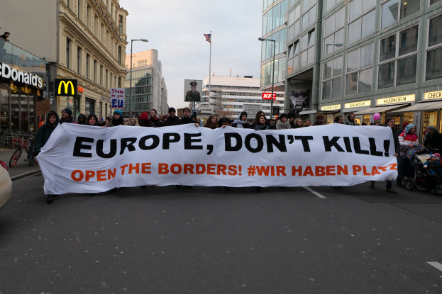 Eine Gruppe von Menschen marschiert auf der Straße entlang und hält ein Banner in Händen, auf dem "Europa, töte nicht öffne die Grenzen" steht, mit einem geparkten Auto links, Gebäuden und einer Flagge im Hintergrund unter einem klaren blauen Himmel.