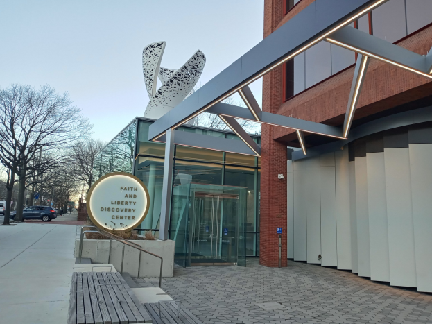 Eingang zum Faith and Liberty Discovery Center mit Glaswänden, einem Namensschild, einer Bank, Metallrahmen, Bäumen, Fahrzeugen, Pfosten und einem bewölkten Himmel.