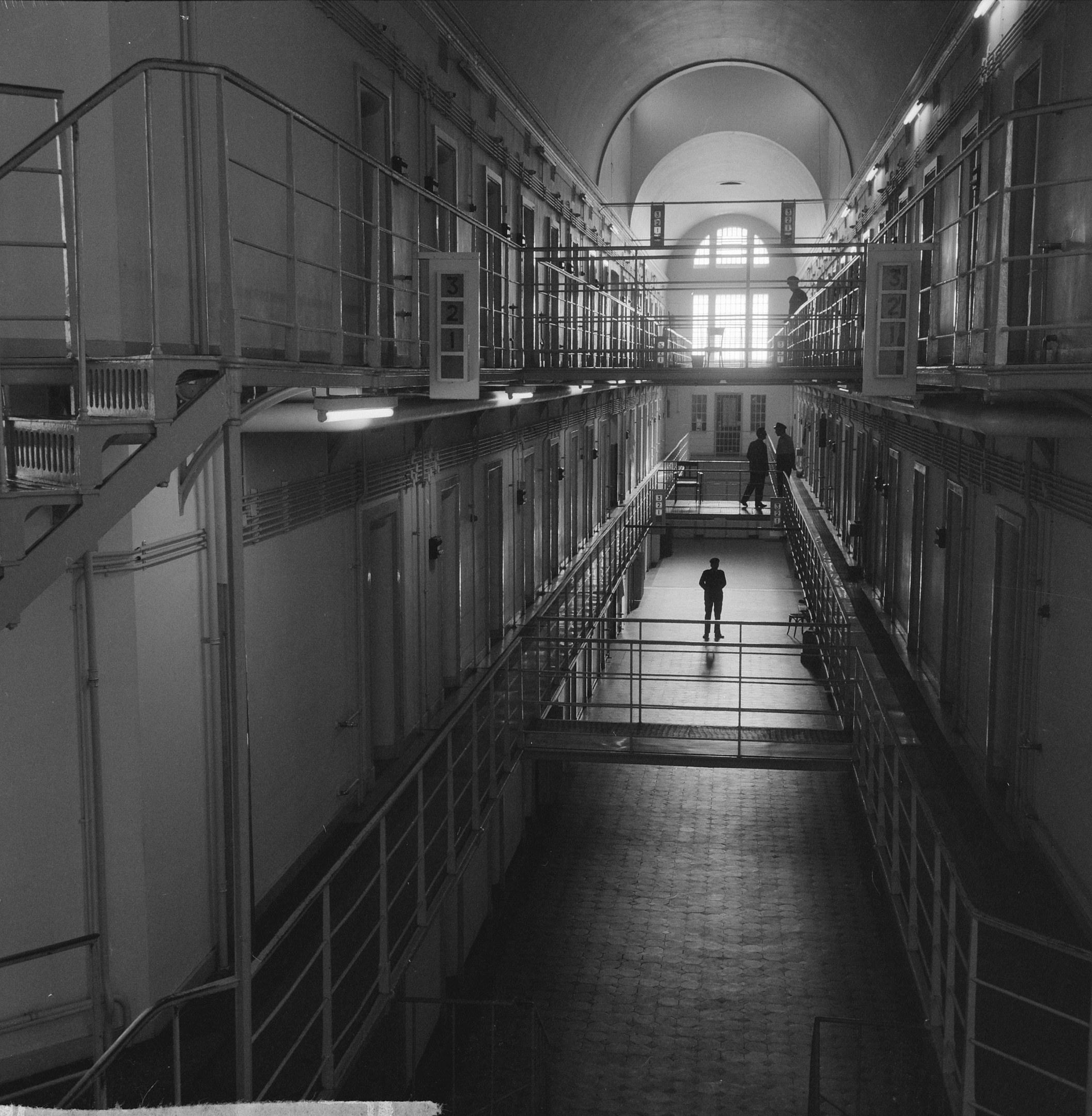 Schwarz-weißes Foto eines Mannes, der einen langen Gefängnishofgang mit Geländern, Türen, Lampen und einem Fenster im Hintergrund entlanggeht.