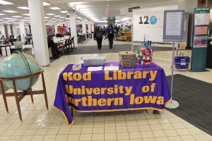 Eine Ausstellung mit einem Tisch, auf dem verschiedene Landesflaggen, eine Box, Papiere und ein Banner liegen, einem Globus links daneben, einer Anschlagtafel rechts daneben mit ein paar Leuten davor und mehreren Tischen und Stühlen links im Hintergrund.