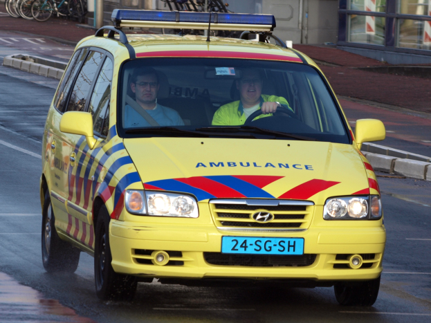 Ein gelber Krankenwagen mit zwei sichtbaren Insassen fahrt an parkenden Fahrr├Ądern auf einem Gehweg vorbei, mit einem Glasfronthaus im Hintergrund.