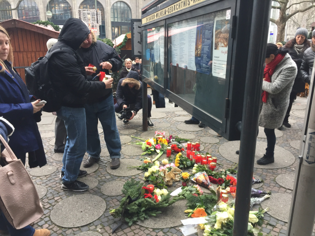 Eine Gruppe von Menschen versammelt sich an einer Bushaltestelle, legt Blumen und Kerzen auf den Boden, einige halten Gegenstände und einer hält eine Kamera; im Hintergrund ist ein Gebäude mit Bögen und Fenstern, Bäume und ein Schuppen zu sehen.