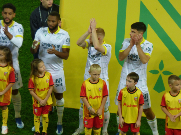Group of soccer players in uniforms clapping on a field with "Ligue 1" sign in the background, celebrating a victory.