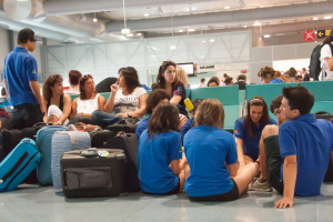 Ein belebter Flughafeninnenraum mit vielen Menschen, die auf Flüge warten, Gepäck auf der linken Seite und Glastüren mit Richtungsschildern im Hintergrund.