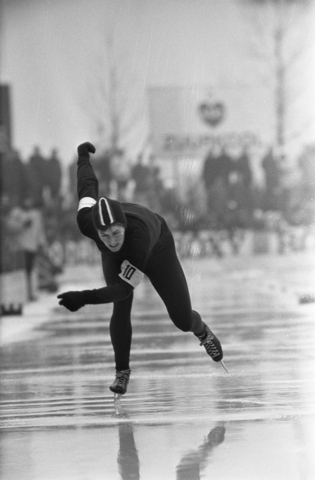 Schwarzes und weißes Foto eines Eisschnellläufers in Bewegung auf dem Eis, der eine Mütze und Rollschuhe trägt, mit unscharfen Figuren, Bäumen und Gebäuden im Hintergrund.