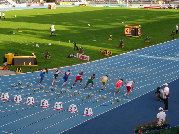 Group of people running on a stadium track with equipment and spectators in the background under a clear blue sky.