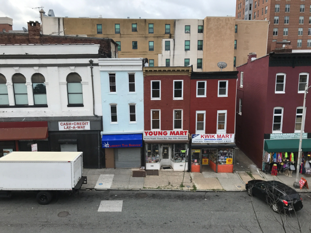 Luftaufnahme einer Stadtstraße in Brooklyn, New York, mit Fahrzeugen, Geschäften, Beschilderungen, Versorgungsmasten, Drähten und einem bewölkten Himmel.