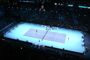 Ein Tennisplatz mit zwei Spielern, Bannern an den Seiten, einer sitzenden Menge im Stadion und Videokameras, mit einem Bild einer Person oben.
