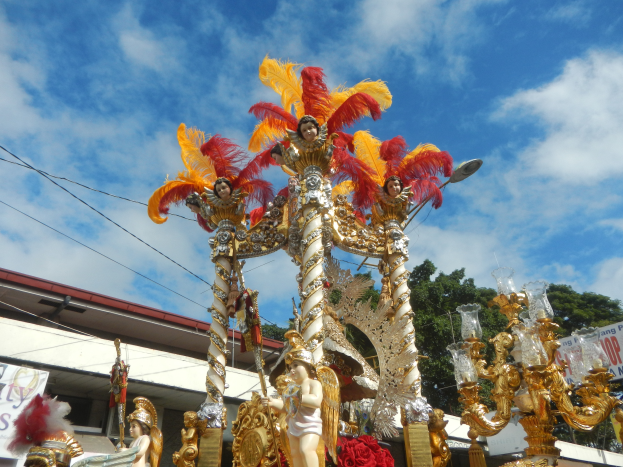 Ein Karnevalswagen, geschmückt mit goldenen und roten Federn, der Menschen trägt, ist in einem Umzug mit einem Gebäude, Bäumen, Drähten und einem Banner in der Nähe zu sehen, unter einem sichtbaren Himmel.
