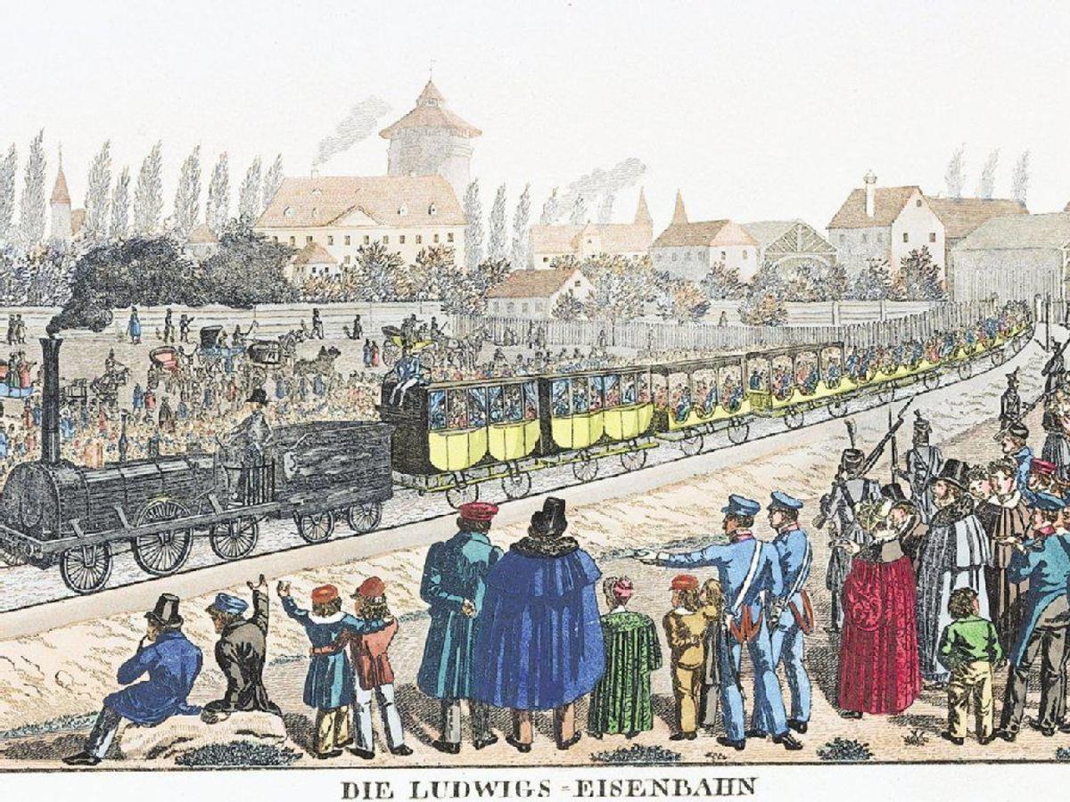 A group of people standing around a train on tracks with some seated inside and others on the ground, set against buildings and trees in the background, with text at the bottom reading "Ludwigs-Eisenbahn, the first railway station in Germany."
