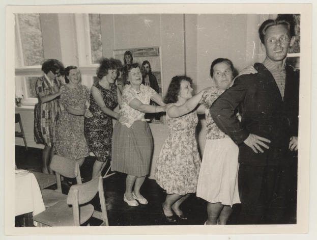 Gruppe von Menschen, die in einem Raum im Stil der 1950er Jahre tanzen, mit Stühlen, einem Tisch mit einer wei├čen Decke, einem Foto auf dem Wand und Fenstern im Hintergrund.
