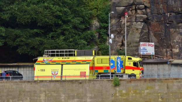 Ambulanz am Straßenrand neben einer Klippe geparkt, mit verschiedenen Straßenobjekten im Vordergrund und Bäumen und einem Auto im Hintergrund.