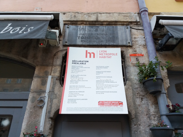 Gebäude mit "Lyon Metropole Habitat"-Schriftzug, einer Tür, einem Anschlagbrett, Topfpflanzen, einem Rohr, Außenbeleuchtung und einem Dach.