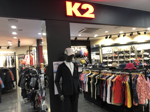 Interior view of a K2 clothing store in Singapore displaying mannequins in caps, hanging clothes, racks of shoes, and illuminated by overhead lights, with a wall-mounted advertisement board.