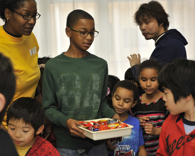 Gruppe von Kindern um eine Frau mit einem Tablett voller Pralinen in einem Raum mit Vorhängen, wahrscheinlich ein Waisenhaus.
