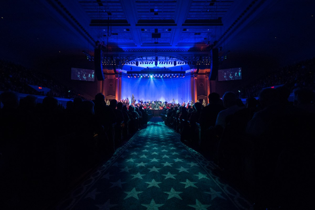 Ein großer Zuschauerraum mit Menschen, die in Reihen vor einer Bühne sitzen, auf der Musiker spielen, unter einer Decke mit Lampen und Fernsehern, was auf einen Konzertsaal der Nationalen Convention 2019 hinweist.