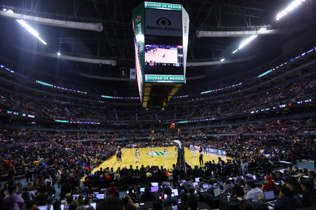 Ein Basketballspiel in einer großen Arena mit Zuschauern auf Sitzen und Stehplätzen, Tischen mit Laptops und Flaschen und einem Basketballkorb, einer Anzeigetafel und Deckenlampen im Hintergrund.