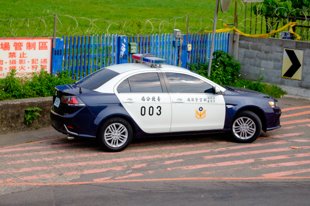 Ein Polizeiwagen am Straßenrand geparkt, umgeben von verschiedenen Gegenständen wie Pflanzen, einem Zaun, einer Texttafel, einer Wand, einem Pfahl, einer Schüsselantenne und Bäumen.
