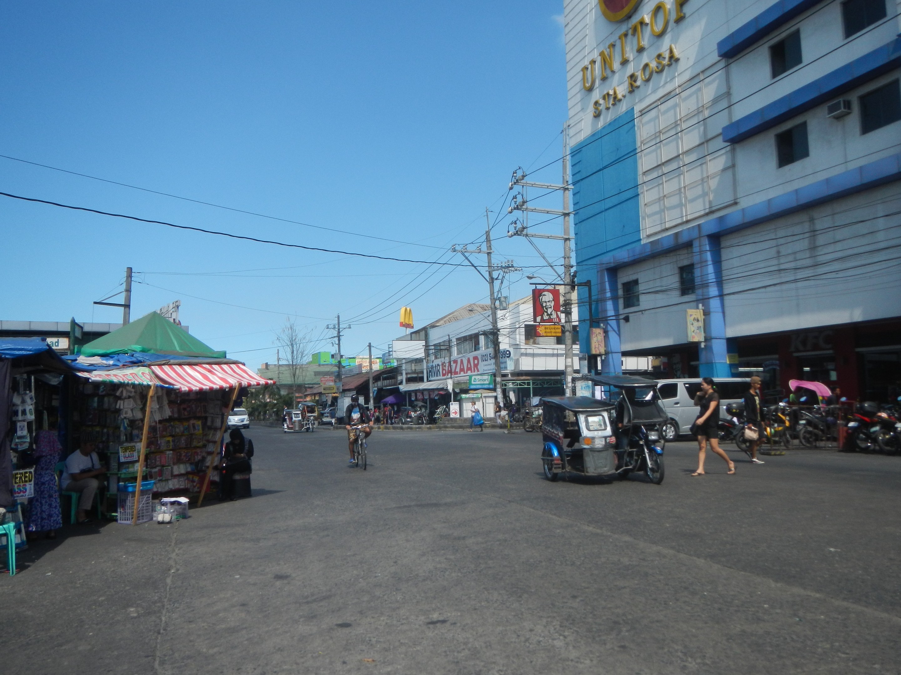 Eine belebte Stadtstraße mit Fahrzeugen, Fußgängern, Marktständen, Gebäuden, Strommasten, Bäumen und einem klaren Himmel.
