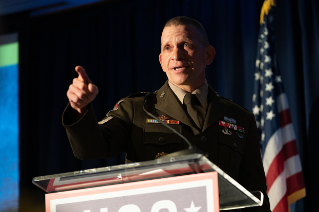Mann in militärischer Uniform, der vor einem Rednerpult mit Mikrofon und einer Tafel mit Text spricht. Im Hintergrund ist ein Vorhang und eine Flagge zu sehen, sowie ein Bildschirm links von ihm.