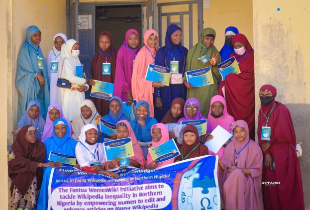 Gruppe von Frauen, die strahlen und Zertifikate in der Hand halten, vor einem Gebäude mit einem Banner, auf dem "Funtua Women Wiki Initiative möchte die Wikipedia-Ungleichheit im nördlichen Nigeria angehen" steht.