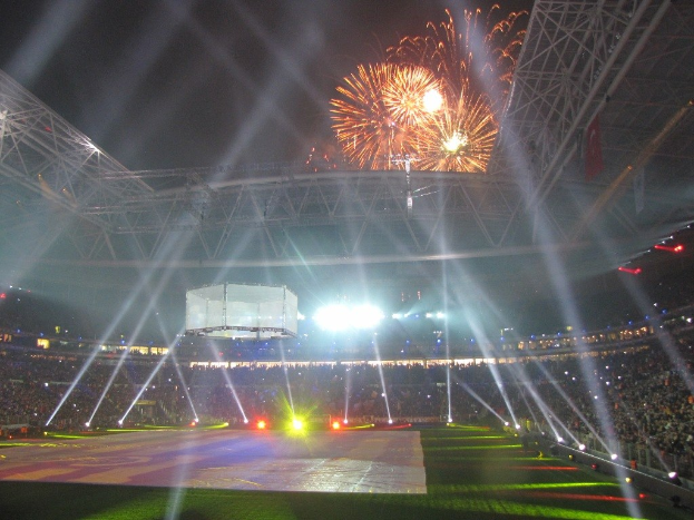 Olympisches Stadion in London bei Nacht, hell erleuchtet mit grünem Rasen, umgeben von einer großen Menge und Feuerwerk, das den Himmel erhellt.