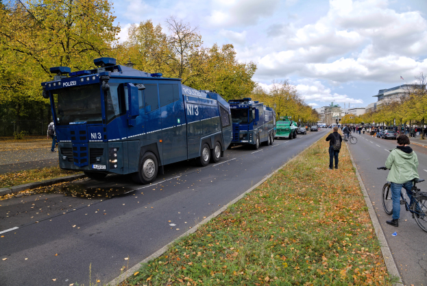 Eine Gruppe von Polizeiwagen, die an einer grasbewachsenen Straße geparkt sind, mit einem Radfahrer auf der rechten Seite, umgeben von Menschen mit Fahrrädern, Bäumen, Gebäuden und einem bewölkten Himmel.