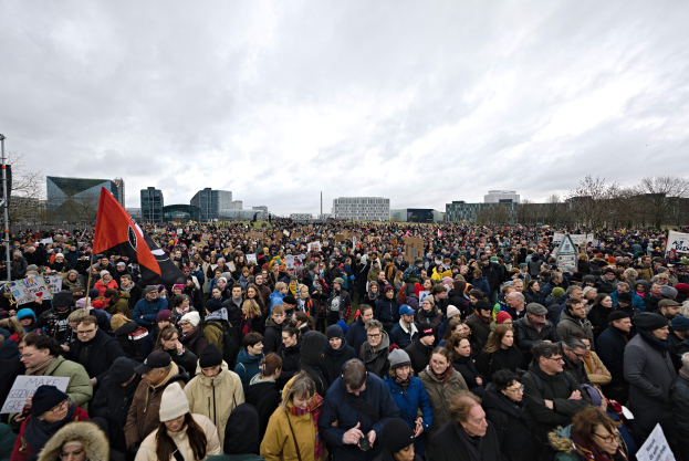 Eine große Menschenmenge steht vor einem Gebäude und hält Schilder und eine Fahne hoch, mit Bäumen und Pfählen im Hintergrund und einem klaren Himmel darüber, wahrscheinlich bei einer Klimaprotestveranstaltung.
