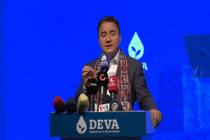 Ein Mann in einem Mantel und Schal steht an einem Podium mit Mikrofonen und spricht zu einem Publikum. Das Podium trägt ein Schild mit der Aufschrift "deva-Geschäftsführer sagt, es sei keine gute Idee für die Zukunft". Im Hintergrund gibt es ein Objekt und einen Bildschirm.