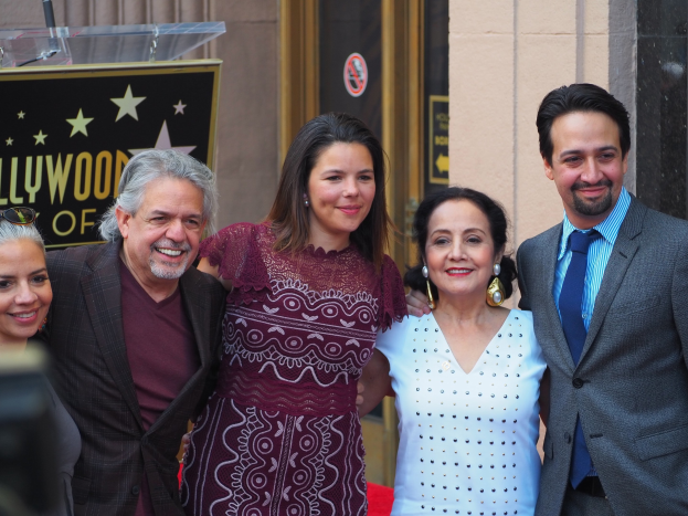 Gruppe von Menschen auf einem roten Teppich bei der Zeremonie der Hollywood Walk of Fame mit einer Tafel, einem Glaspodium und einer Wand im Hintergrund und einer Kamera auf der linken Seite.