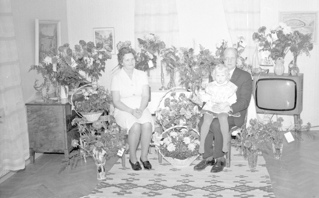 Schwarz-weiß-Foto einer Familie aus drei Personen - einem Mann, einer Frau und einem Kind - die auf einem Sofa sitzen, mit Blumenvasen auf benachbarten Tischen, einem Teppich auf dem Boden, Vorhängen im Hintergrund und Fotorahmen an der Wand.