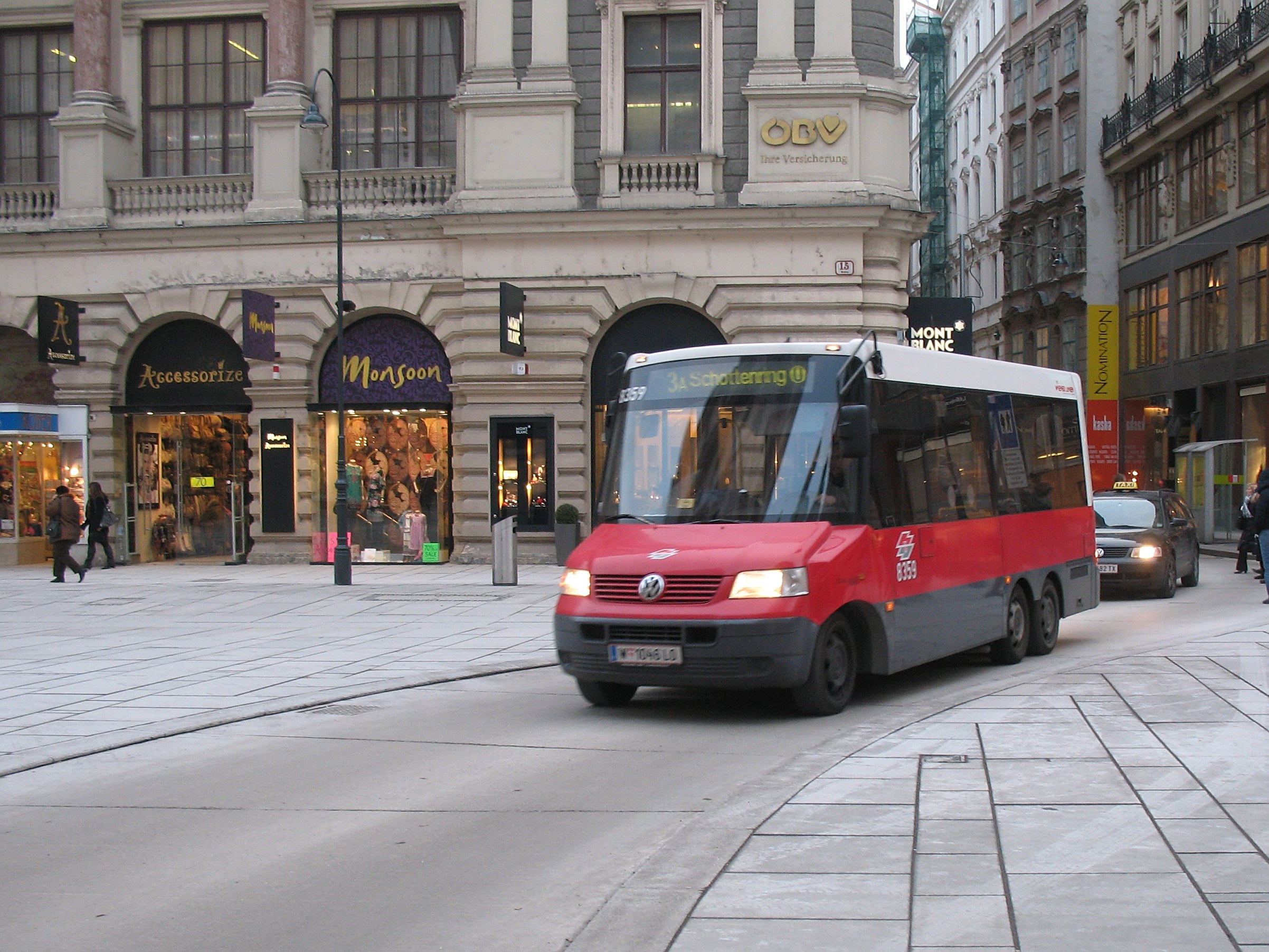 A red shuttle bus drives down a city street lined with tall buildings, pedestrians on the sidewalk, light poles, and stores with visible name boards under a visible sky.