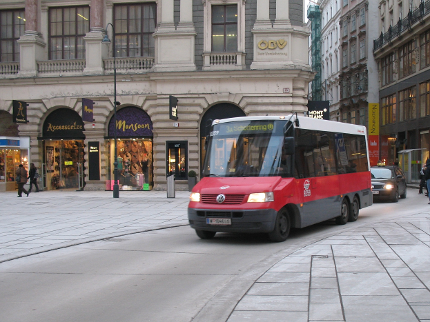 A red shuttle bus drives down a city street lined with tall buildings, pedestrians on the sidewalk, light poles, and stores with visible name boards under a visible sky.