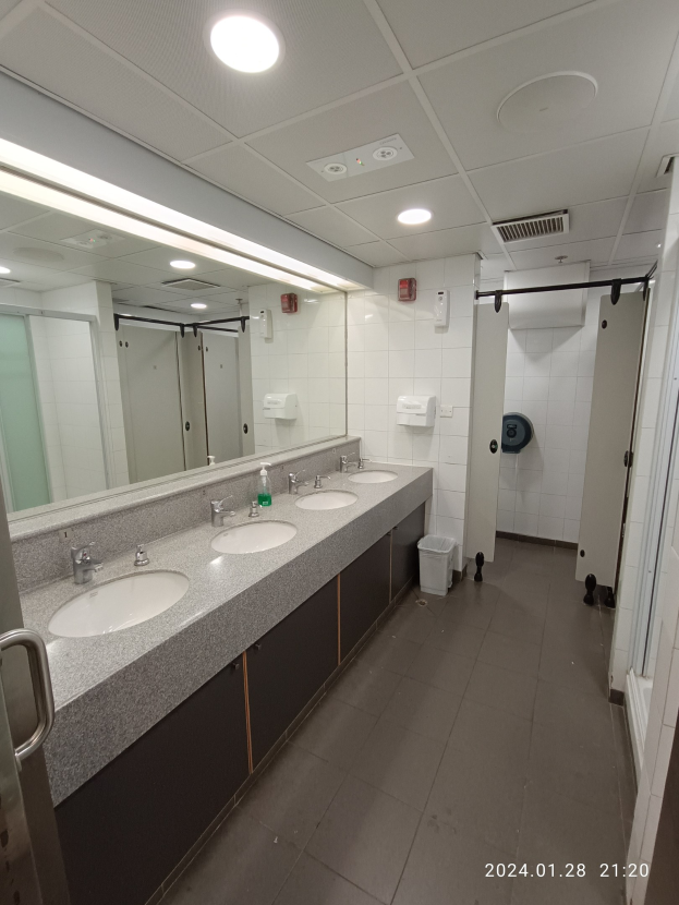 Öffentliche Toilette mit zwei Waschbecken, einem großen Spiegel, Wasserhähnen, einem Bodenmülleimer, einer Tür mit Griff und Deckenleuchten.