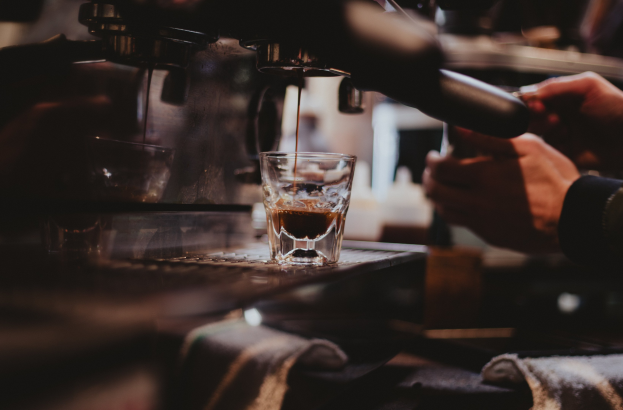 Ein Barista bereitet einen Kaffee an einer Kaffeemaschine zu, mit einem Glas Kaffee und einem drapierten Tuch auf dem Tisch, Hintergrund leicht unscharf.