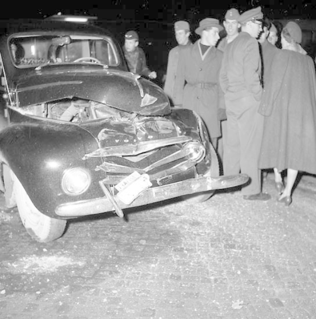 Schwarz-weißes Bild eines stark beschädigten Autos am Straßenrand mit einer Gruppe von Menschen, die es inspizieren.